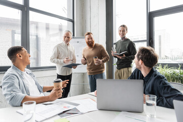 Cheerful business people talking to interracial colleagues with clipboard during training in office.