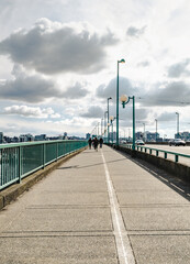 Fototapeta premium Back view of pedestrian crossing bridge with city background on a sunny spring day. Long bridge with shared sidewalk and vehicle traffic lanes. Cambie Bridge, Vancouver, BC, Canada. Selective focus.