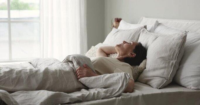 Carefree peaceful mature woman waking up on orthopedic mattress, resting in white bedding, sitting up, stretching body, rising hands, enjoying good morning after healthy sleeping