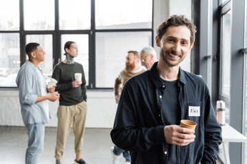 Smiling man holding paper cup and looking at camera during alcoholics meeting in rehab center.