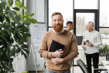 Smiling businessman holding notebook near blurred plants and multiethnic colleagues in office.