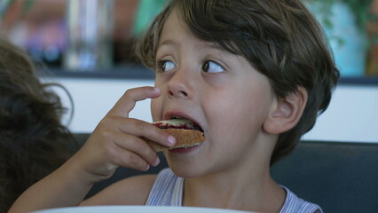 Child eating bread with jelly in morning breafast table. Candid small boy takes a bite of food