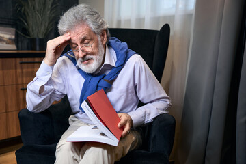 Bearded old male sits in a cozy armchair with book