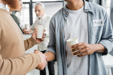 Obraz na płótnie Canvas Interracial men with paper cups shaking hands during alcoholics meeting in rehab center.