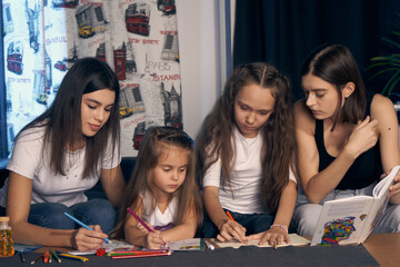 Staged photo. Lesbian couple and their kids are having a good time at home.  Parents and girls sitting at the table with pencils. The little girl watches with interest as her older sister draws.