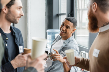 Obraz premium Cheerful african american man holding thermos and talking to people during alcoholics meeting in rehab center.
