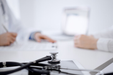 Stethoscope lying on the tablet computer in front of a doctor and patient sitting opposite each other at the background . Medicine, healthcare concept.