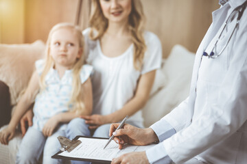 Fototapeta premium Doctor and patient. Pediatrician using clipboard while examining little girl with her mother at home. Sick and unhappy child at medical exam