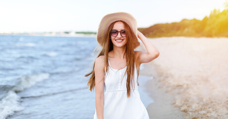 Fototapeta na wymiar Happy smiling woman in free happiness bliss on ocean beach standing and posing with hat and sunglasses. Portrait of a female model in white summer dress enjoying nature during travel holidays vacation