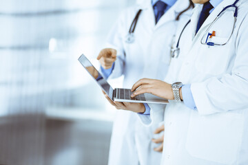 Two unknown doctors with stethoscopes discuss medical exam resoults, while standing at hospital office. Physicians using a laptop for checking up medication names records