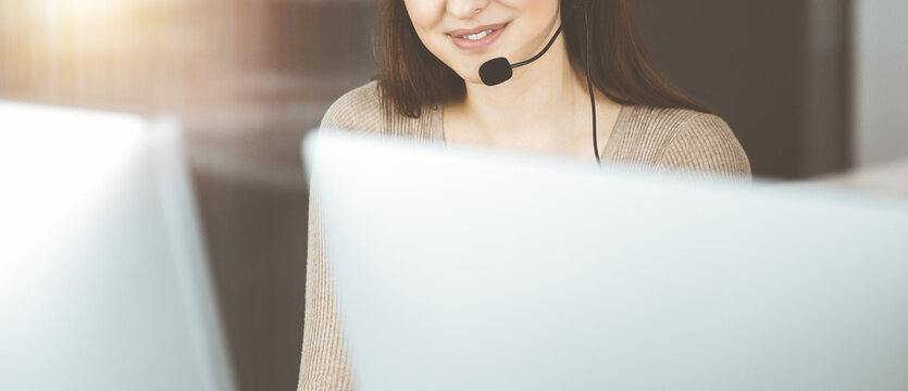 Young girl in headsets is talking to a firm's client, while sitting at the desk in sunny office.