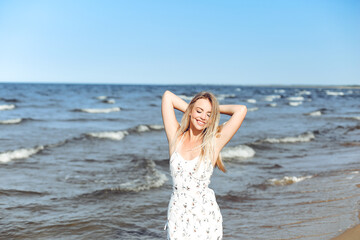 Fototapeta na wymiar Happy blonde beautiful woman on the ocean beach standing in a white summer dress, raising hands.