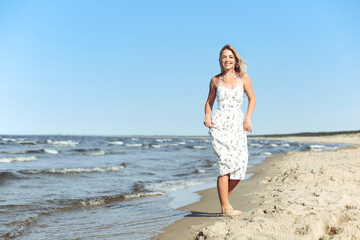 Happy blonde woman in free happiness bliss on ocean beach standing straight. Portrait of a female model in white summer dress enjoying nature during travel holidays vacation outdoors