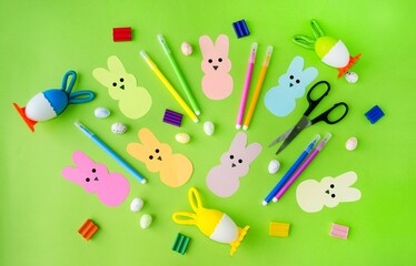 On a green background, Easter pepps bunnies cut out of colored paper, felt-tip pens, colored eggs, scissors, plasticine.  Flat lay, top view.  Concept craft, applique, children's creativity.