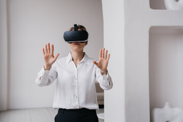 Young female student in white shirt and black pants using vr glasses exploring arts and museums standing indoors against wall with ceramics  on shelves. Girl using virtual reality. Education, game.