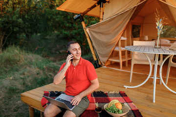 Happy businessman freelance using laptop while talking by phone on cozy glamping tent in a summer day. Luxury camping tent for outdoor holiday and vacation. Lifestyle concept