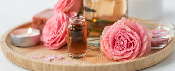 Aromatherapy. Pure organic essential rose oil concept. Elixir with plant based floral herbal ingredients. Pink flowers extract. Spa atmosphere with candle, towel. White background. Banner copy space