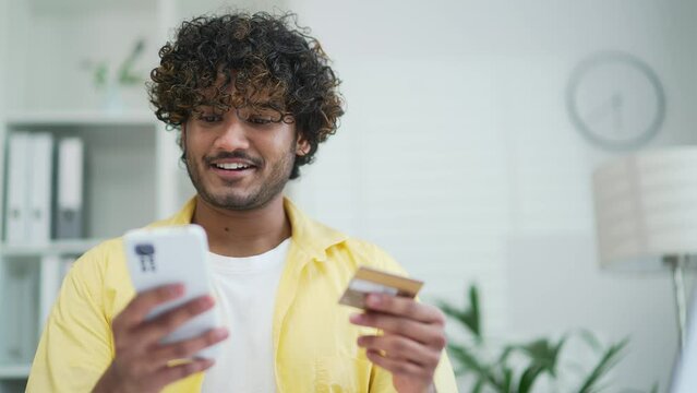 Young adult man pay for services makes a payment using smartphone and credit card, sitting at office or home Happy male using a mobile app booking orders by phone Makes purchases in online stores