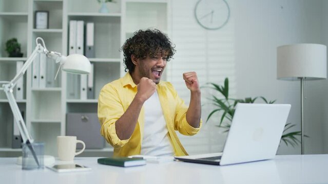 Excited young fan freelancer employee looking at laptop computer screen watch online sport game cheering man celebrating win goal rejoices victory good news at office workplace success great result