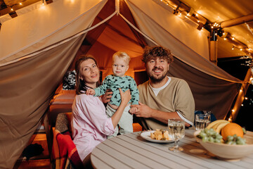 Happy family with lovely baby have dinner and spend time together in glamping on summer evening near cozy bonfire. Luxury camping tent for outdoor recreation and recreation. Lifestyle concept