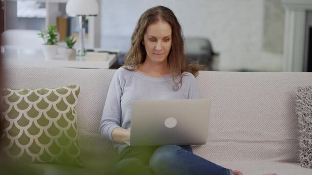 Woman at home, sitting on couch in living room, working with laptop computer in home office. Portrait of happy adult caucasian woman in 30s, smiling.