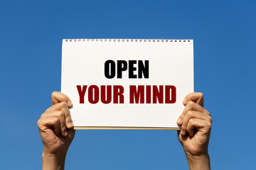 Open your mind text on notebook paper held by 2 hands with isolated blue sky background. This message can be used as business concept about opening your mind.