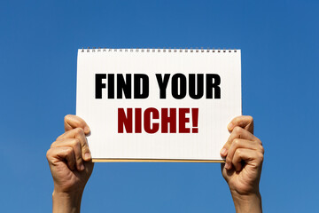 Find your niche text on notebook paper held by 2 hands with isolated blue sky background. This message can be used as business concept about finding your niche.