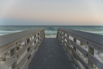 Wooden path heading to the beach shore against the horizon sky in Destin, Florida. Straight walkway above grassy sand dunes against the view of ocean waves under sunset skyline.