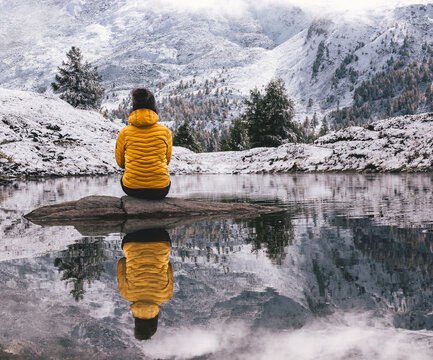 person in yellow jacket reflection in a calm pond in the mountains with snowy mountains in distance