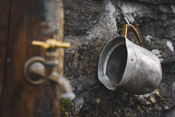 Old aluminum jug hanging on the wall next to a water tap - 579874305