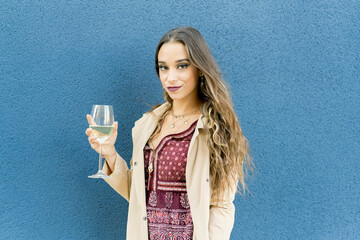 Young smiling female in stylish wear with ornament and makeup standing with glass of white wine while looking at camera