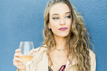 Young woman in stylish wear with ornament and makeup standing with glass of white wine while looking away