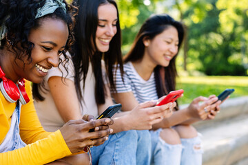 Three joyful multiracial young teenage girls having fun together outdoors. Addicted millennial female watching social media content on smart phone device app. Technology lifestyle concept