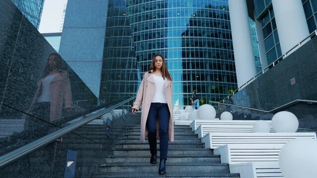 Slim girl with long hair quickly descends stairs in urban street. Young woman in raincoat going down steps from tall modern building. Beautiful lady is in hurry on business in metropolis. Front view