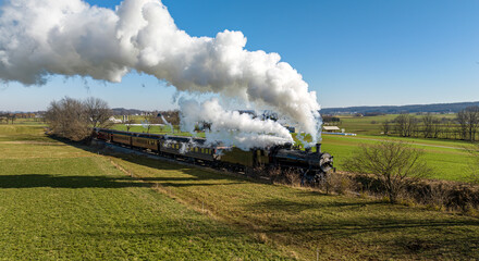 Obraz na płótnie Canvas A Drone View of an Antique Steam Passenger Train Approaching Traveling Thru Countryside and Farmland Blowing Smoke on a Sunny Winter Day