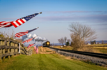Fototapeta premium A View of a Classic Steam Passenger Train Approaching, With American Flags Attached to a Fence on a Sunny Summer Day