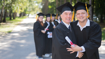 A group of graduates in robes outdoors. An elderly man and a young guy congratulate each other on...