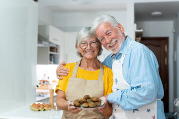 Headshot portrait of cute old couple of seniors showing to camera what they cooked. Together people at home cooking in the kitchen. Healthy food concept diet holding meatballs.