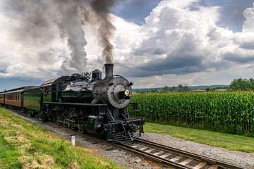 Fototapeta na wymiar A Steam Passenger Train Passing Corn Fields and Blowing Smoke on a Sunny Summer Day