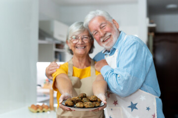 Headshot portrait of cute old couple of seniors showing to camera what they cooked. Together people at home cooking in the kitchen. Healthy food concept diet holding meatballs.