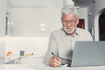 Fototapeta premium Aged remote worker. Concentrated senior male in glasses work on laptop from home office read email electronic document. Old age man employee freelancer sit at kitchen table by pc typing report online.