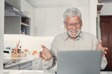 Fototapeta premium Elderly grey haired man sit at table wear glasses looks at laptop screen talk via video call app. Older patient receive on-line counselling, virtual meeting of old friends or family concept.