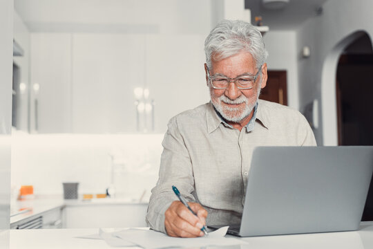 Aged remote worker. Concentrated senior male in glasses work on laptop from home office read email electronic document. Old age man employee freelancer sit at kitchen table by pc typing report online.