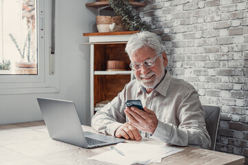 Old man in glasses sit in kitchen near laptop holding smartphone device chat remotely, browse webpage, use new mobile app enjoy usage of modern wireless tech, make business from home distantly concept