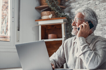 Portrait of smiling mature man sit at table at home talk on cellphone work on computer gadget....