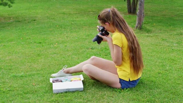 Female food photographer sits on grass, takes pictures of donuts inside paper packaging. Girl shooting doughnuts with professional camera in box on lawn for her blog about nutrition, diets, fast food