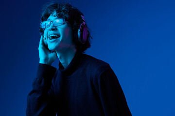Teenage man wearing headphones listening to music and dancing and singing with glasses, hipster lifestyle, portrait blue background, neon light, style and trends, mixed light, copy space