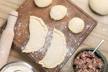 Raw chebureki with tasty filling and meat in bowl on wooden table, flat lay