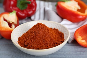Bowl with aromatic paprika powder and fresh bell peppers on grey wooden table, closeup