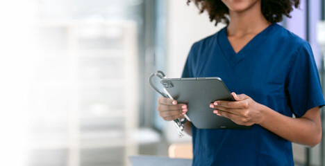 Fototapeta premium Black female nurse or doctor holding stethoscope and digital tablet standing in hospital office, copy space.